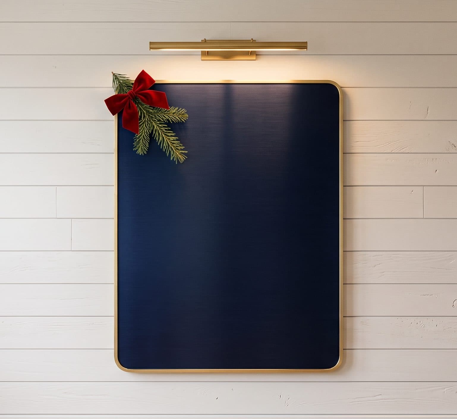 A navy plaque with a gold border on a white shiplap wall under a brass picture light, with an evergreen sprig and red bow at its corner